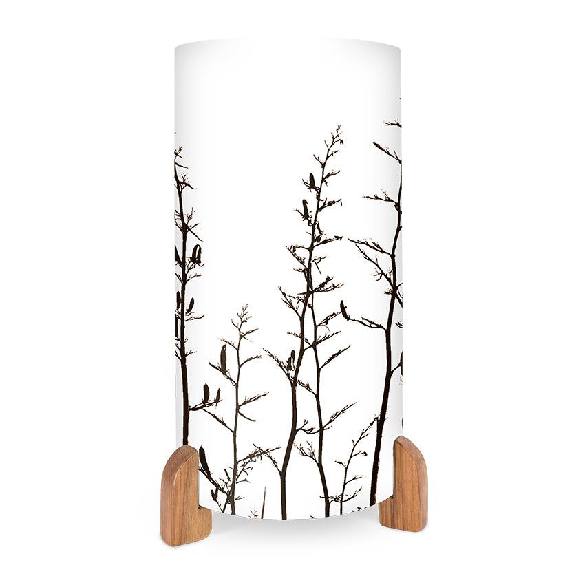 The Harakeke Table Lamp is a cylindrical white lamp with black tree branches and birds, featuring an in-line switch and two wooden base feet.