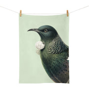 The Hushed Green Tui Tea Towel, designed in New Zealand and screen printed with a dark green and white bird illustration, hangs from two wooden clothespins against a light background.