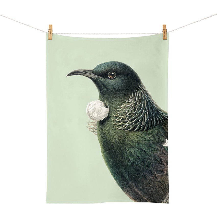 The Hushed Green Tui Tea Towel, designed in New Zealand and screen printed with a dark green and white bird illustration, hangs from two wooden clothespins against a light background.