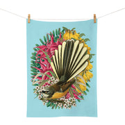 The Botanical Fantail Tea Towel, clipped to a clothesline, showcases a New Zealand fantail with a fanned tail among colorful flowers and green foliage on a light blue background.