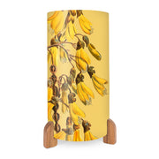 The Vintage Botanical Kowhai Table Lamp features wooden feet and a curved yellow shade adorned with vibrant kowhai flower and brown seed pod images.