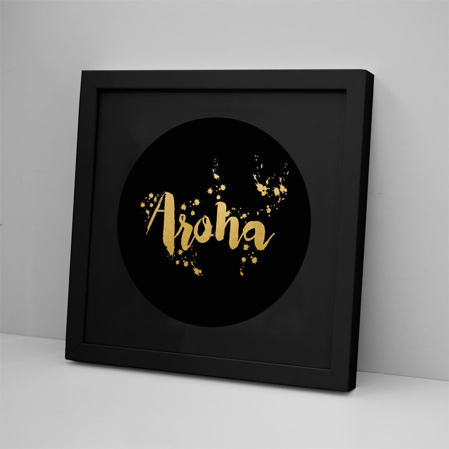 The Aroha Foil Print features a black circle and gold-scripted “Aroha” with gold splatters, available framed or unframed. Set on a white surface against a light gray wall, its black frame complements the Kiwiana-inspired art.