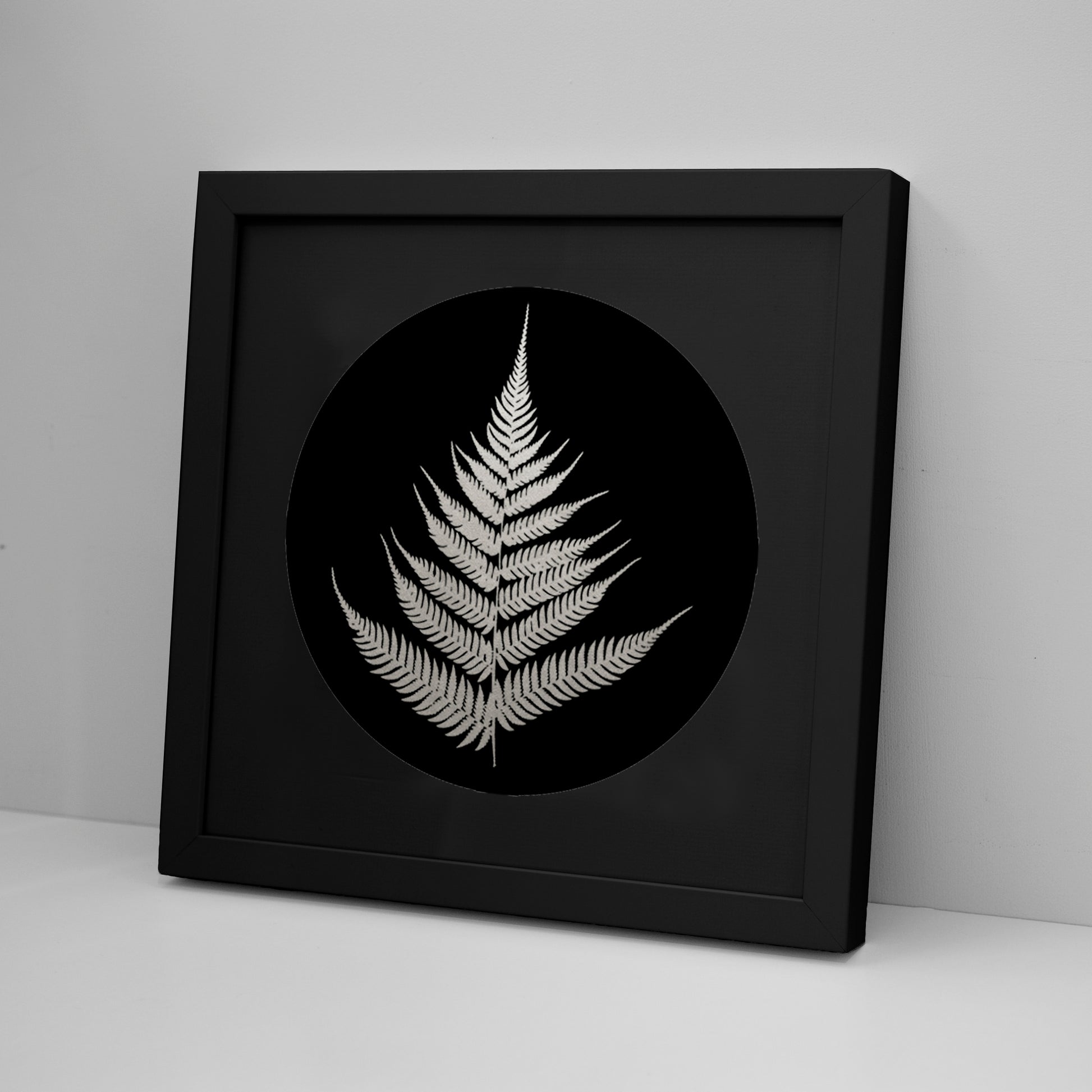 The Silver Fern Foil Print displays a detailed white fern leaf on a black circular background, inspired by Kiwiana. Choose framed or unframed options to suit your space.