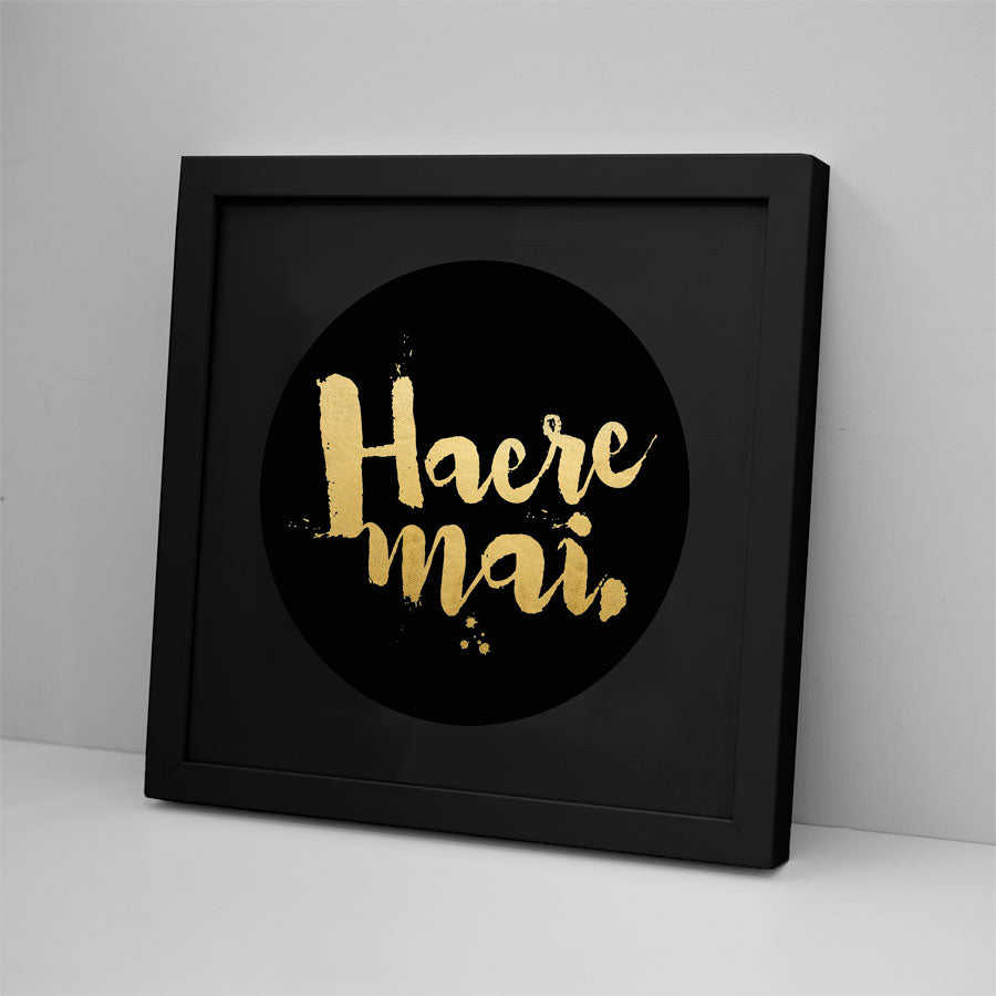 The Haere Mai - Foil Print features gold script on a black circular background, set in a black frame for a bold look, and is shown against a light gray wall.