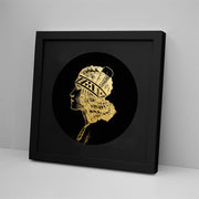 The Wahine Foil Print features a gold foil profile of a person with a feathered headdress on a black circular background, displayed in a sleek black frame resting on a modern white surface against a plain wall.