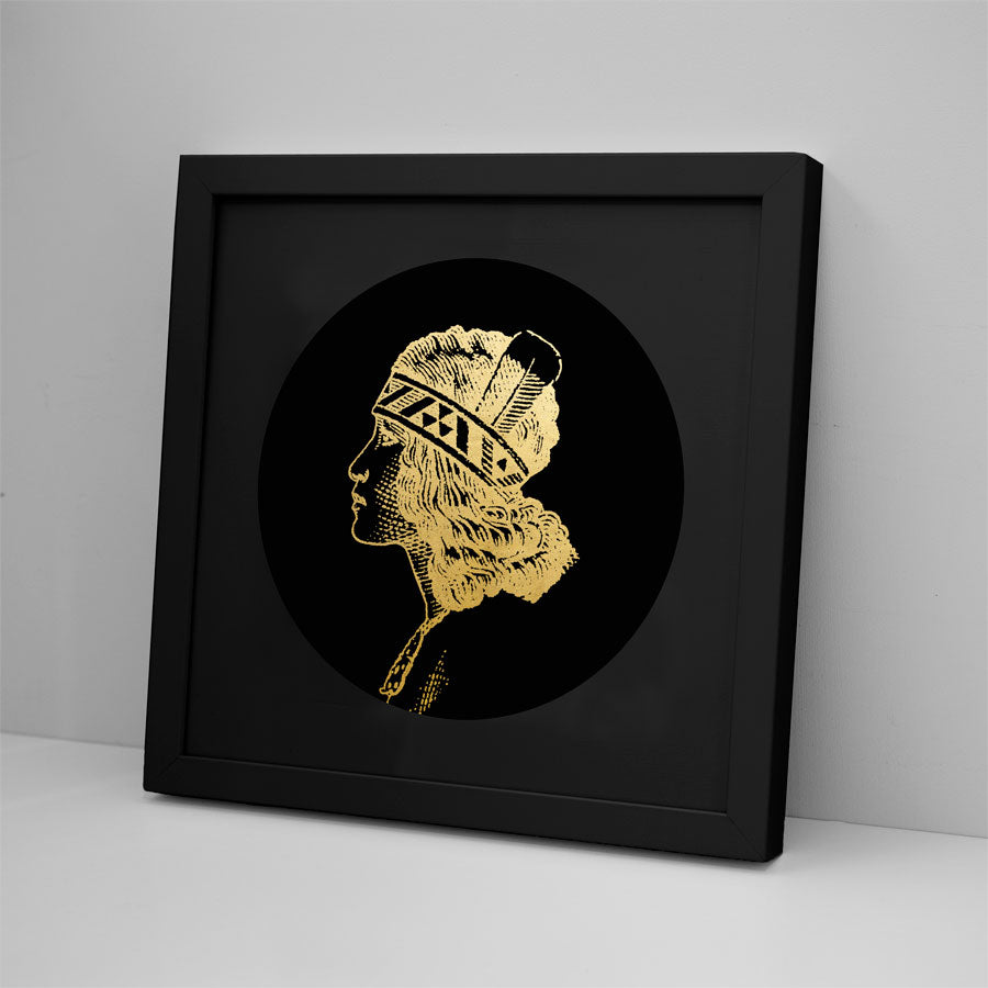 The Wahine Foil Print features a gold foil profile of a person with a feathered headdress on a black circular background, displayed in a sleek black frame resting on a modern white surface against a plain wall.
