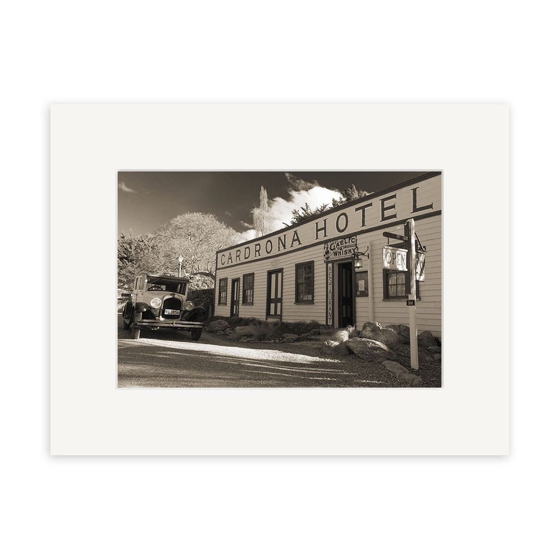 A sepia photo features the historic Cardrona Pub - RP071 in New Zealand, with a vintage car, wooden façade, hotel and garage signs, and rocky landscaping captured in stunning detail for photographic prints.