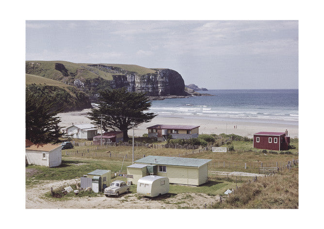 Retro Bach Print captures small cabins and a caravan by Jacks Bay’s sandy beach, set against grassy hills and cliffs, with a few people strolling along the shore beneath a partly cloudy sky. Archival quality photographic print.