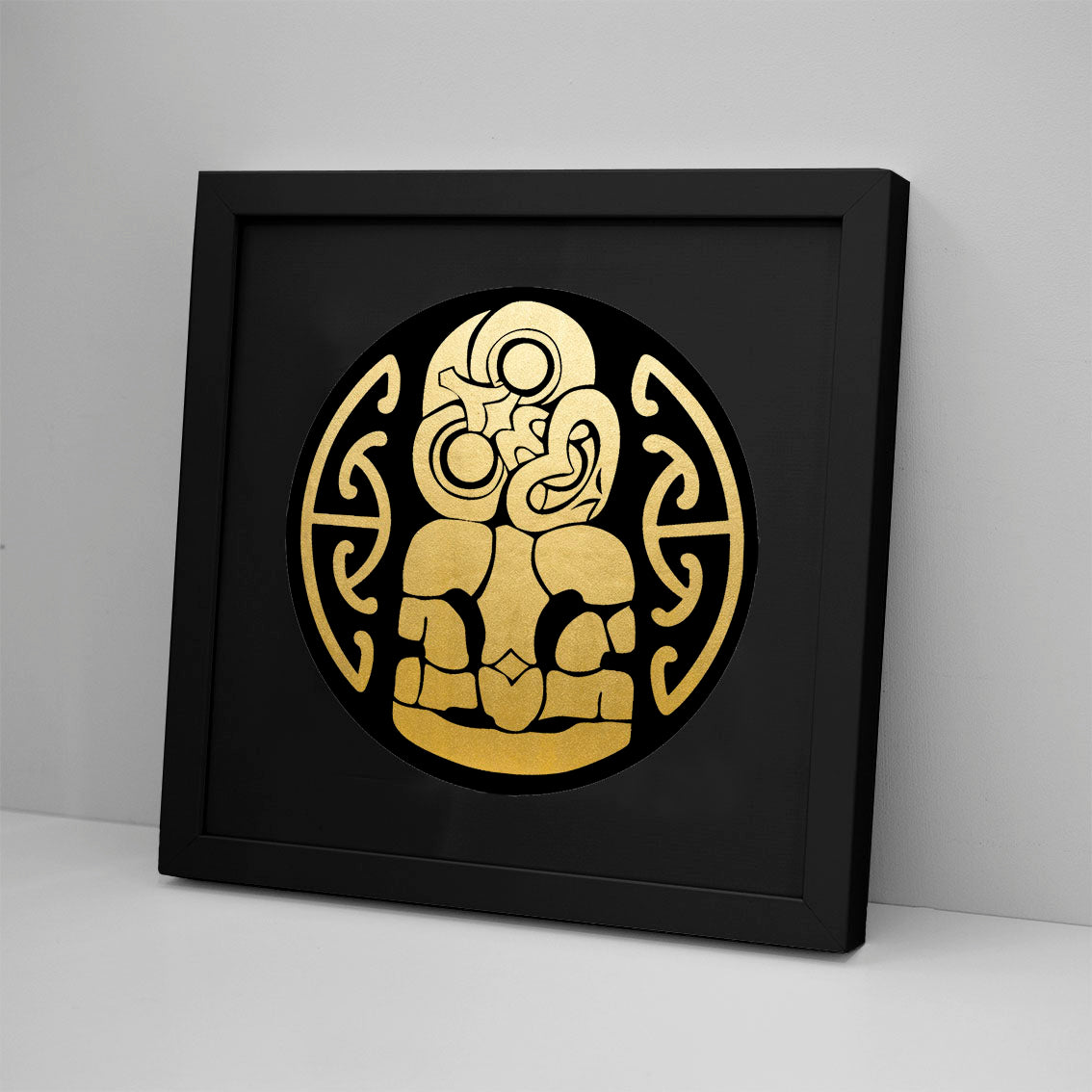 The Tiki Foil Print is a framed artwork with a circular gold design on black, featuring abstract, symmetrical patterns in curved and angular shapes. This gold foil print, made in New Zealand, leans against a light-colored wall.