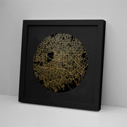 The Christchurch Mapscape Foil Print features a black frame and a circular gold-foiled map of city streets on a black background, shown leaning against a white wall on a flat white surface.