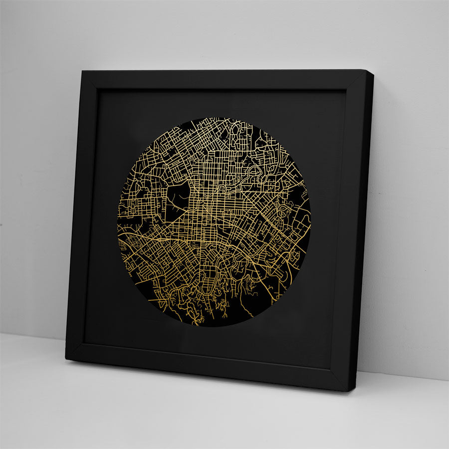 The Christchurch Mapscape Foil Print features a black frame and a circular gold-foiled map of city streets on a black background, shown leaning against a white wall on a flat white surface.